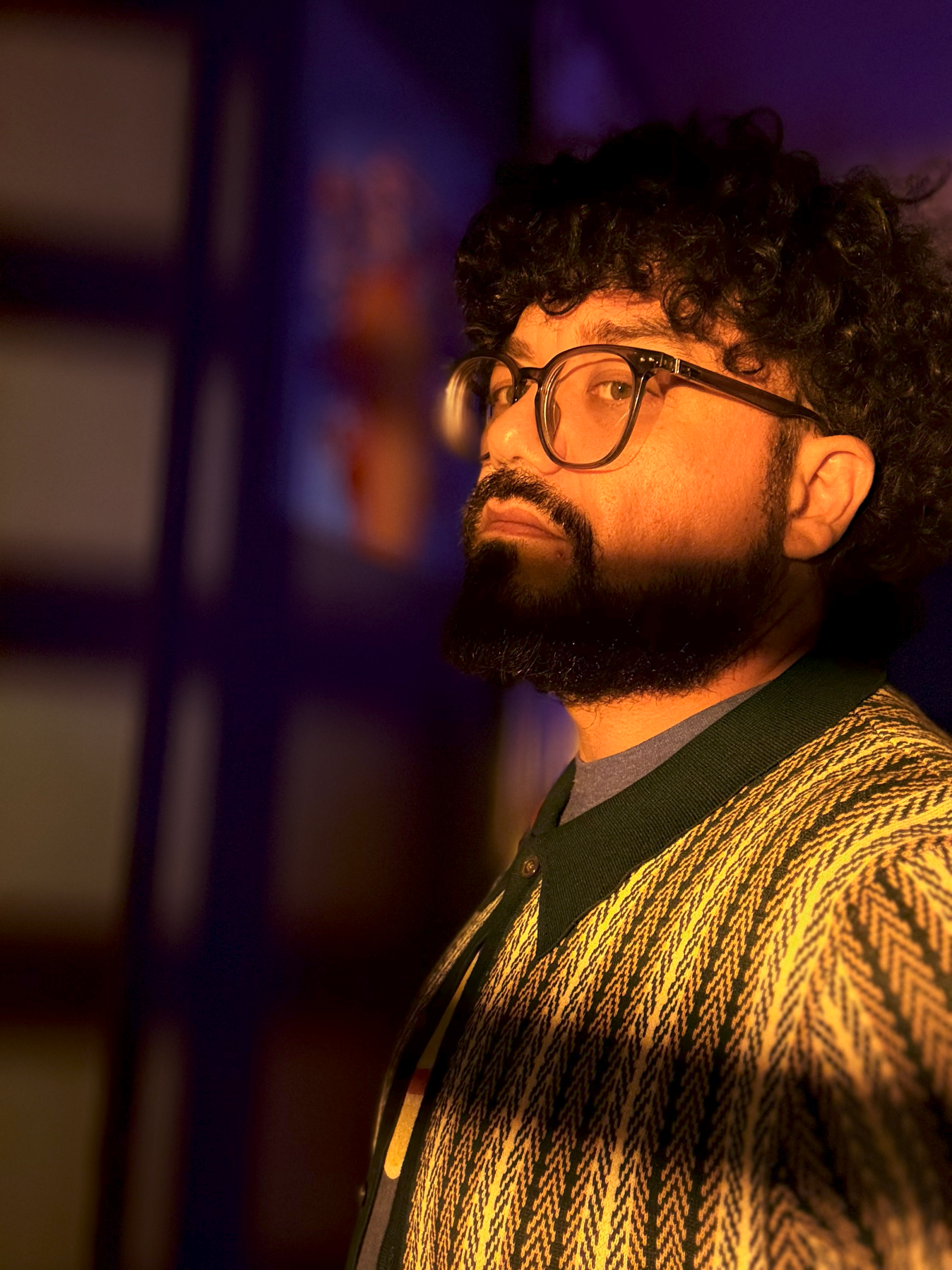 Side profile portrait of a person with curly hair, glasses, and a beard, wearing a patterned sweater and lit with warm, moody lighting.